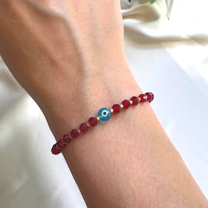 Puede incluir: Una pulsera de cuentas rojas con un dije de ojo turco turquesa. La pulsera está hecha de pequeñas cuentas rojas redondas y espaciadores dorados. El dije de ojo turco es de color turquesa brillante con una pupila negra.