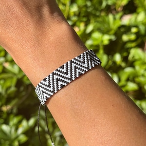Puede incluir: Pulsera con patrón de chevrón en blanco y negro. La pulsera tejida presenta un diseño en zigzag repetido. La pulsera es ajustable con cordones negros y pequeñas cuentas blancas. La pulsera se muestra en una muñeca.
