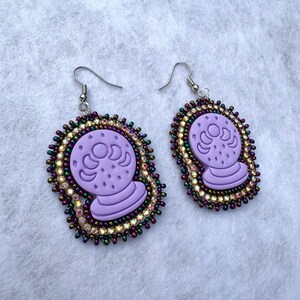 May include: A pair of lavender-colored crystal ball earrings. Each earring features a crystal ball design with crescent moon details, surrounded by iridescent and gold-toned beads. The earrings have silver-colored fish hook closures.