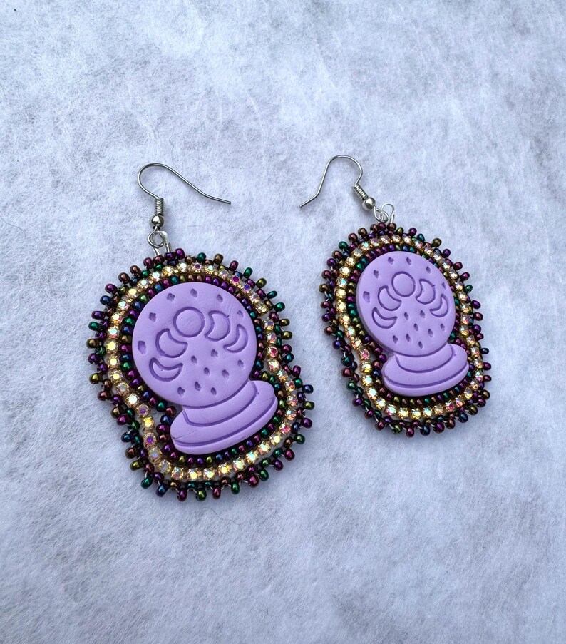 May include: A pair of lavender-colored crystal ball earrings. Each earring features a crystal ball design with crescent moon details, surrounded by iridescent and gold-toned beads. The earrings have silver-colored fish hook closures.