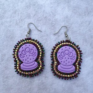 May include: A pair of purple crystal ball earrings. Each earring features a lavender crystal ball design with a beaded border of iridescent and gold beads. The earrings have silver-colored fish hook ear wires.