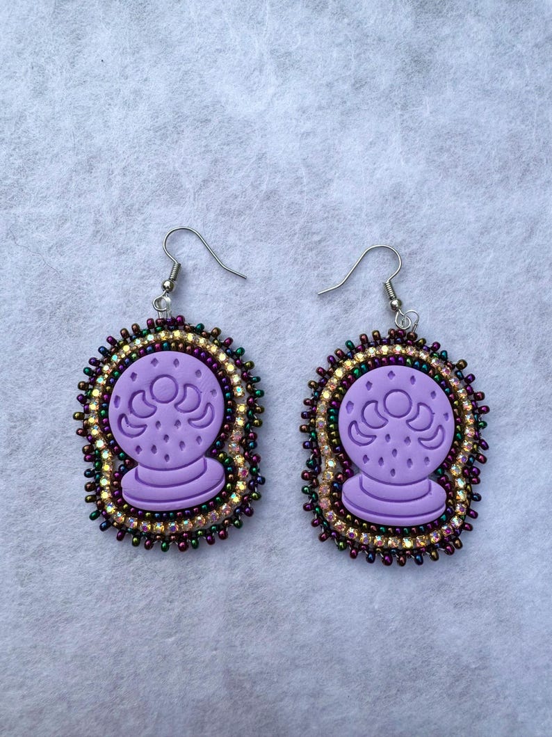 May include: A pair of purple crystal ball earrings. Each earring features a lavender crystal ball design with a beaded border of iridescent and gold beads. The earrings have silver-colored fish hook ear wires.