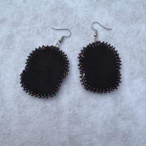 May include: A pair of black felt earrings with a beaded edge. The earrings have a teardrop shape and are outlined with small, iridescent beads in shades of purple, green, and gold. The earring hooks are silver.