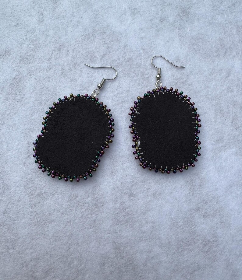May include: A pair of black felt earrings with a beaded edge. The earrings have a teardrop shape and are outlined with small, iridescent beads in shades of purple, green, and gold. The earring hooks are silver.