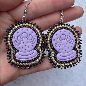 May include: A pair of lavender-colored earrings featuring a crystal ball design. The earrings have a beaded border with iridescent and purple beads. The crystal ball design is embossed on the lavender base, and the earrings have silver-colored ear wires.