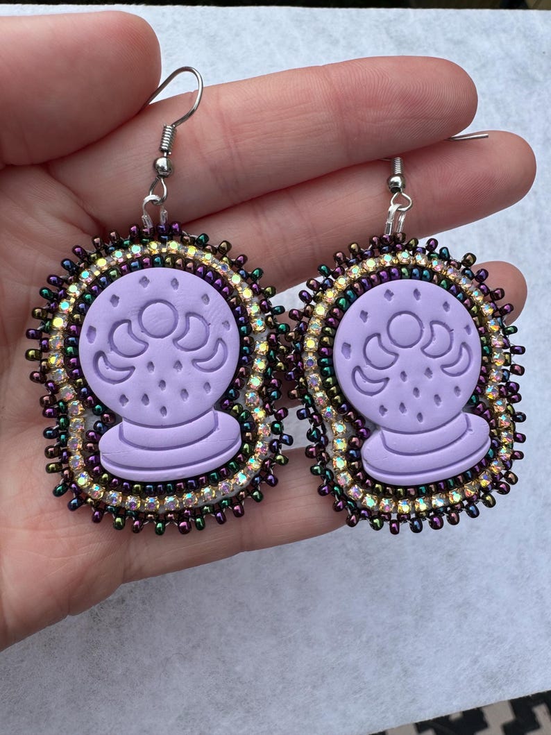 May include: A pair of lavender-colored earrings featuring a crystal ball design. The earrings have a beaded border with iridescent and purple beads. The crystal ball design is embossed on the lavender base, and the earrings have silver-colored ear wires.