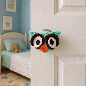 May include: A crochet owl door knob cover featuring a bright turquoise body, large dark brown and white circular eyes with black pupils, and a small orange triangular beak. The cover is fitted over a round metal door knob on a white door.
