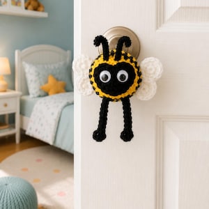 May include: A handmade crochet bee door knob cover. The bee has a yellow and black body, googly eyes, white wings, and black antennae and legs. The door knob cover is attached to a white door.