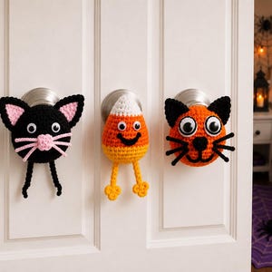 May include: Three crochet door knob covers featuring a black cat with pink accents, a candy corn character with a smiling face, and an orange cat with black ears and whiskers. Each piece has googly eyes and hangs from a silver door knob.