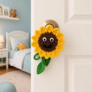 May include: A handmade crochet sunflower door hanger with a smiling face and googly eyes. The sunflower has yellow petals, a brown center, and green leaves. It is attached to a white door with a brass doorknob.