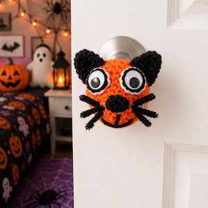 May include: A handmade orange and black crocheted cat face door knob cover with large googly eyes and black whiskers. The cat face is attached to a silver door knob on a white door. Halloween decor.