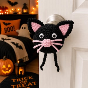 May include: A black crocheted cat face door knob cover with pink inner ears, a pink nose, and white whiskers. The cat has large googly eyes and dangling black legs. The door knob cover is a Halloween decoration.