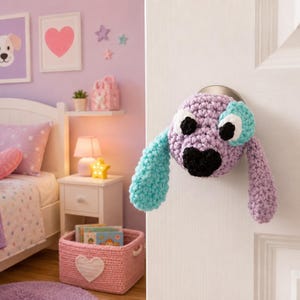 May include: A crochet dog door knob cover featuring a light purple head, a black nose, large white and black eyes, and two long floppy ears in contrasting light purple and bright turquoise yarn, attached to a round metal door handle.