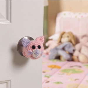 May include: A pink and lavender crocheted cat head door knob cover with black button eyes and light pink whiskers. The cat is attached to a silver door knob on a white door. The background is a blurred bedroom scene.