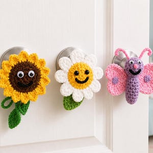 May include: Three crochet door knob covers featuring a yellow sunflower with a brown center, a white daisy with a yellow center, and a purple butterfly with pink wings. Each piece includes googly eyes and a stitched black smile.