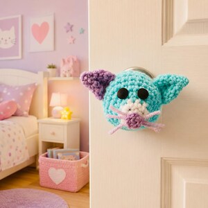May include: A turquoise crocheted animal cover for a round door knob, featuring two black button eyes, a white muzzle, a small purple nose, purple-tipped ears, and light purple yarn whiskers attached to a standard interior door handle.