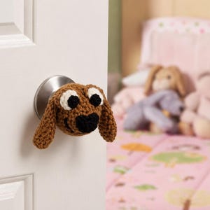 May include: A brown crocheted dog door knob cover with white eyes and a black nose. The cover is attached to a silver door knob on a white door. The background is a blurred image of a pink bed with stuffed animals.
