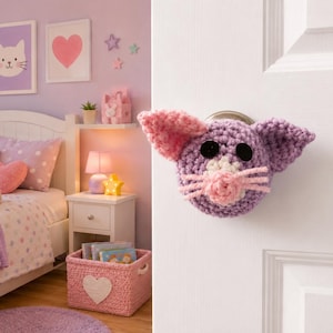 May include: A crochet door knob cover shaped like a cat head, featuring a purple face, pink ears, a white muzzle, pink yarn whiskers, and two black circular eyes. The cover is fitted over a silver-toned metal door knob on a white door.