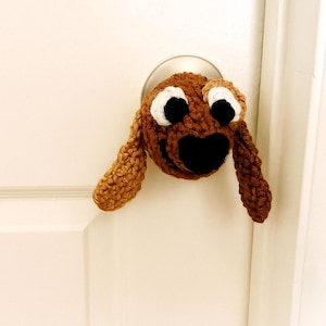 May include: A handmade, brown crocheted dog head door knob cover with white accents around the eyes and a black nose. The dog's ears hang down on either side. The door knob cover is attached to a white door.