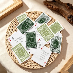 Op de afbeelding: Een verzameling speelkaarten met aangepaste groen-witte, bloemrijke achterkantontwerpen met namen en datums, naast kaarten bedrukt met tekst waaronder "Wishing you both a lifetime of joy" en "May your life be filled with warmth and gentle surprises."