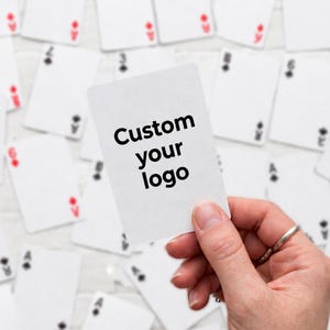 Peut inclure: Une carte à jouer blanche avec le texte "Custom your logo" en noir est tenue devant un fond éparpillé de cartes à jouer. Les cartes en arrière-plan ont des enseignes rouges et noires.