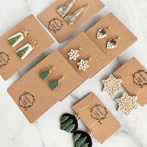 Puede incluir: Una colección de pendientes navideños hechos a mano, presentados en tarjetas marrones. Los pendientes presentan diseños de árboles de Navidad, copos de nieve y adornos, en verde, blanco, dorado y negro. Las tarjetas tienen el logo "Velvet Fern Clay Company".