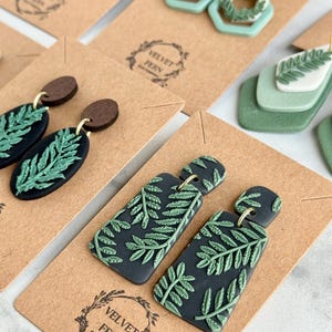 Puede incluir: Varios pendientes de arcilla hechos a mano con diseños de hojas de helecho. Los pendientes son de diferentes formas y colores, incluyendo negro, verde y marrón. Algunos pendientes tienen un acento de madera. Los pendientes se muestran en tarjetas marrones con el logotipo "Velvet Fern Clay Company".