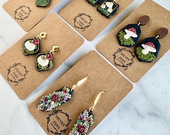 May include: A selection of handmade polymer clay earrings on brown cards. The earrings showcase designs like mushrooms, flowers, and foliage, with gold-coloured hooks. The brand name "Velvet Fern Clay Company" is printed on the cards.