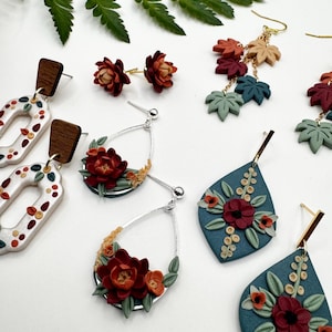 Puede incluir: Una colección de pendientes de arcilla polimérica con diversos diseños florales y de hojas en colores otoñales, incluyendo rojo intenso, verde salvia y naranja apagado. El conjunto incluye estilos de botón, lágrima y colgantes.