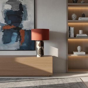 May include: An interior shot showing a wooden cabinet, an abstract painting, and a table lamp. The lamp has a red lampshade and a patterned base. A bookshelf with decorative items and books is also visible.