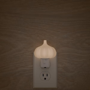 May include: A white night light shaped like a garlic clove is plugged into a white electrical outlet. The night light is illuminated, casting a soft glow. The background is a wooden wall.