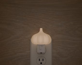 May include: A white night light shaped like a garlic clove is plugged into a white electrical outlet. The night light is illuminated, casting a soft glow. The background is a wooden wall.