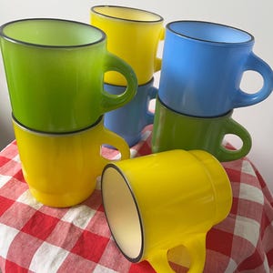 May include: A collection of colorful mugs in shades of green, yellow, and blue. Each mug has a rounded handle and a black rim. The mugs are stacked on top of each other, with one mug lying on its side on a red and white checkered tablecloth.