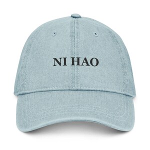 May include: Light blue denim baseball cap with the text "NI HAO" embroidered in black. The cap has a curved brim and an adjustable strap at the back. A casual and stylish accessory.
