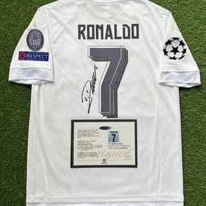 May include: White soccer jersey with the name "RONALDO" and the number "7" in gray. The jersey has a signature and patches on the sleeves. A framed certificate is displayed below the number.