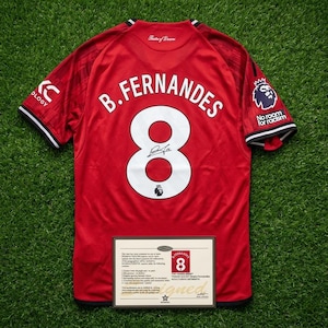 Puede incluir: Una camiseta de fútbol roja con el nombre "B. Fernandes" y el número "8" en blanco. La camiseta tiene una firma en el número y las palabras "No room for racism" en la manga. Se muestra un certificado de autenticidad debajo.