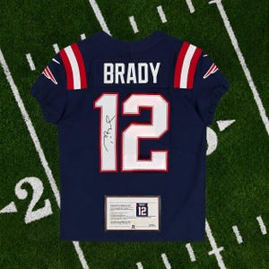 May include: Navy blue football jersey with the name "BRADY" and the number "12" in white. The jersey has red and white accents on the shoulders and a signature on the front. A certificate of authenticity is displayed at the bottom.