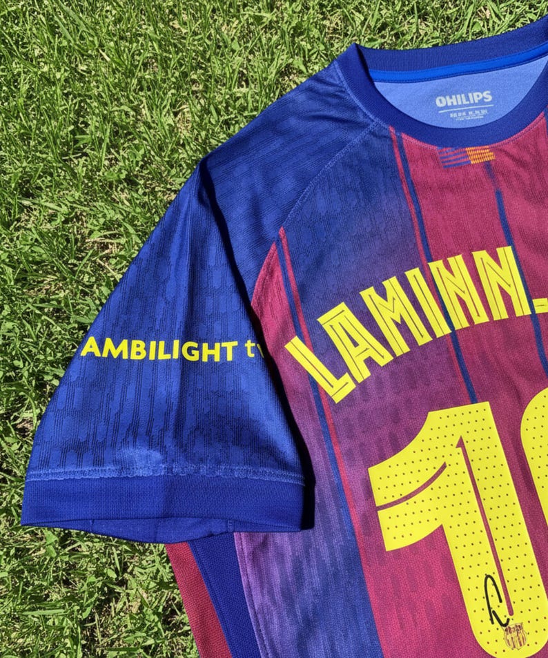 May include: A blue and maroon soccer jersey with the name "LAMINN" and the number "10" in yellow. The sleeve has the text "AMBILIGHT t" in yellow. The jersey is on a green grassy surface.