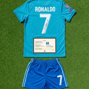 Puede incluir: Un conjunto de camiseta y pantalón corto de fútbol azul claro. La camiseta presenta el nombre "RONALDO" y el número "7" en blanco, junto con una firma. El pantalón corto también tiene el número "7". Se muestra un certificado debajo.