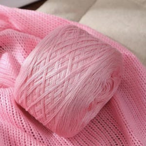 May include: A ball of light pink yarn rests on a matching pink knitted fabric. The yarn is tightly wound, displaying a crisscross pattern. The soft texture of the yarn and fabric suggests a craft project or textile creation.
