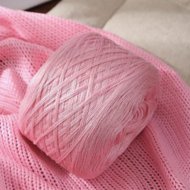 May include: A ball of light pink yarn rests on a matching pink knitted fabric. The yarn is tightly wound, displaying a crisscross pattern. The soft texture of the yarn and fabric suggests a craft project or textile creation.