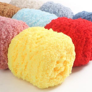 Puede incluir: Una colección de bolas de hilo esponjosas y redondas en varios colores, incluyendo amarillo, rojo, rosa, azul y marrón. El hilo tiene un aspecto suave y texturizado, lo que sugiere una calidad acogedora y táctil. Las bolas están dispuestas juntas.