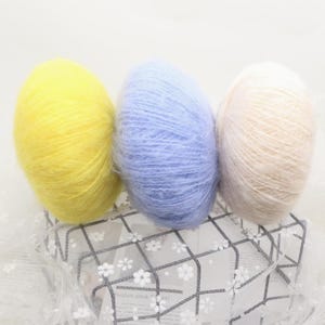 May include: Three balls of soft yarn in yellow, pale blue, and cream. The yarn has a fluffy texture, ideal for knitting or crochet. The colours are arranged together, suggesting they are part of a set or collection for craft projects.