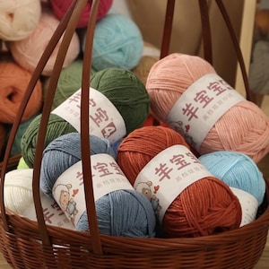 May include: A brown wicker basket overflowing with balls of yarn in various colours, including pink, blue, green, and orange. Each ball has a label with text and a sheep cartoon. The yarn is tightly wound, perfect for knitting or crochet.