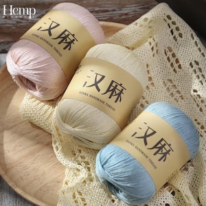 May include: Three balls of yarn in pastel shades: pink, cream, and light blue. Each ball has a brown paper label with Chinese characters and the text "HANMA HANDMADE THREAD". The yarn is presented on a wooden tray with cream-coloured knitted fabric.