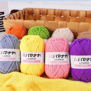 May include: A selection of colourful yarn balls in shades of yellow, pink, purple, and blue, displayed in a woven basket. Each ball has a label with Japanese text and the word "LILIARGE". Ideal for knitting and crochet projects.