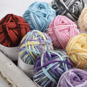 Könnte beinhalten: Eine Sammlung bunter Wollknäuel in verschiedenen Rot-, Blau-, Rosa-, Gelb- und Lilatönen. Die Wolle ist in kleinen Papierbehältern angeordnet, was auf ein Bastel- oder Strickprojekt hindeutet. Das Bild zeigt eine Vielzahl von Texturen und Farbtönen.