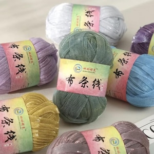May include: A collection of colorful yarn balls in shades of white, lavender, gold, green, blue, and silver. Each ball is wrapped with a label featuring Chinese characters. The yarn has a shiny, metallic appearance.