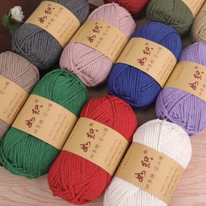 May include: A collection of colorful yarn skeins in various shades, including gray, pink, blue, green, red, and white. Each skein is wrapped with a brown paper label featuring Chinese characters. The yarn is likely made of cotton and polyester.
