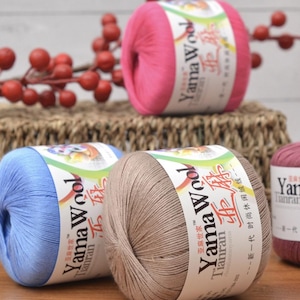 以下が含まれることがあります： ピンク、ブルー、ベージュの色合いの毛糸玉がいくつかあり、「Yama Wool」のラベルが付いています。毛糸は編みかごの上に配置されており、手芸や編み物のテーマを示唆しています。